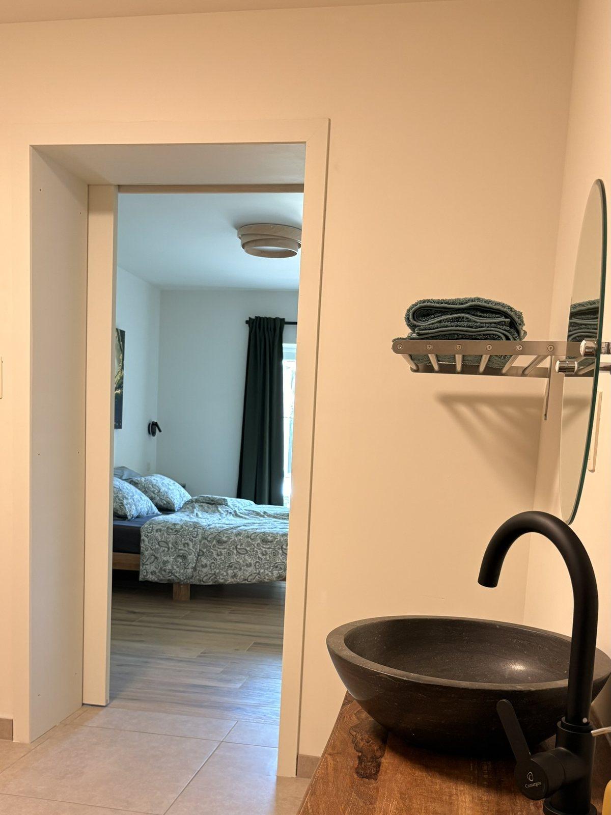 Natuurstenen waskom op houten blad in de badkamer op het gelijkvloers, met doorkijk naar de slaapkamer