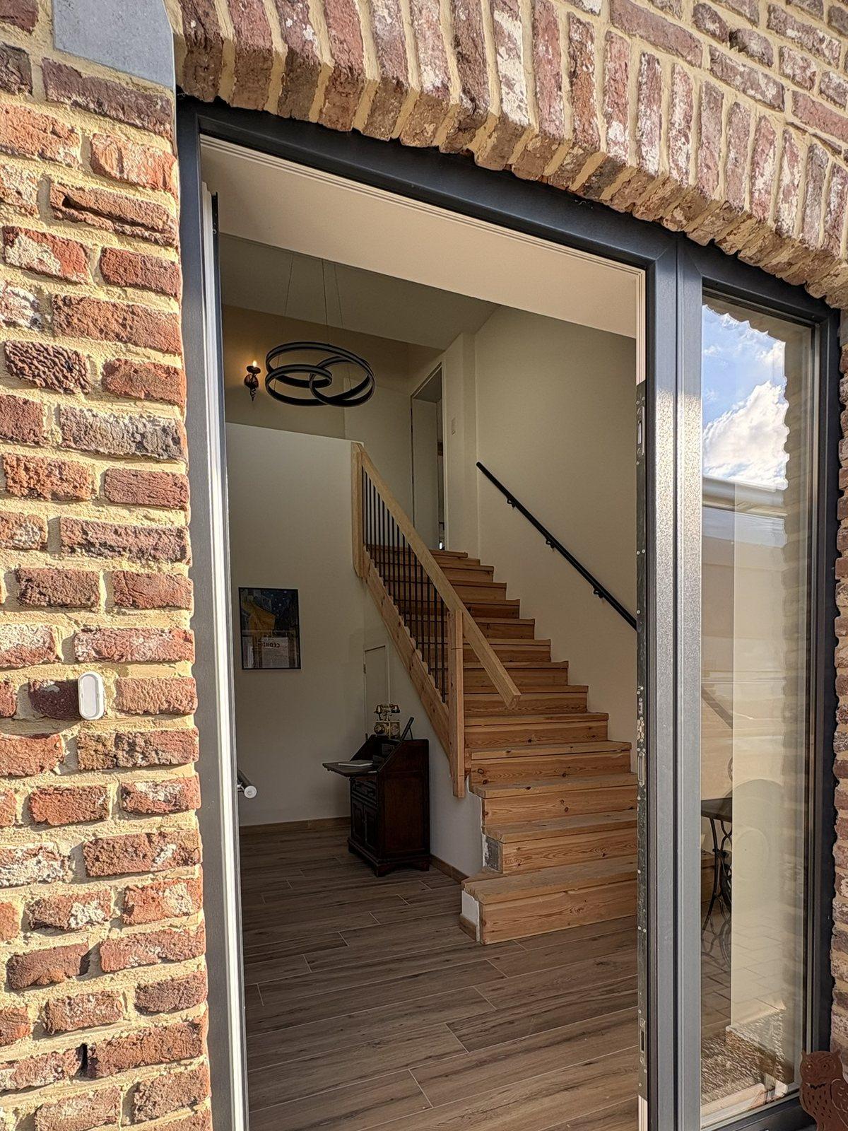 Open voordeur met zicht op de houten trap in de inkomhal van Maison Campagnard, Teuven